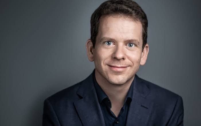 Lächelnder Mann mit kurz geschnittenen Haaren, blauem Rollkragenpullover und weißem Hemd.