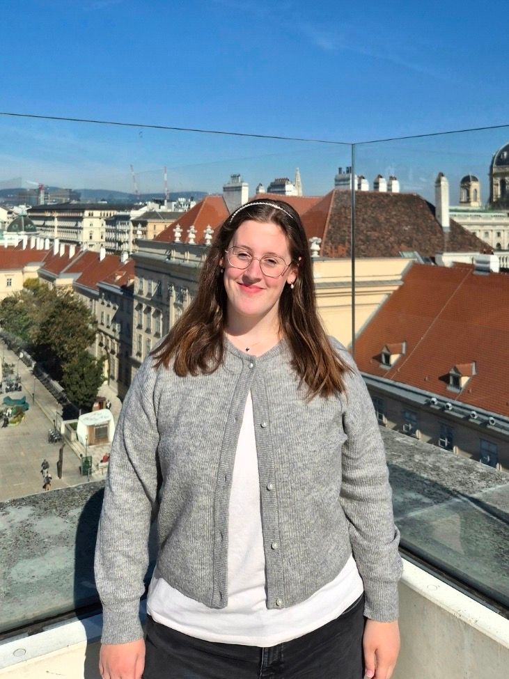 Frau lächelt vor historischer Stadt mit roten Dächern und trägt graue Cardigan-Jacke, Blick in die Ferne.