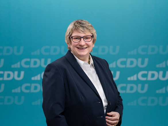 Frau in Anzug vor CDU-Hintergrund lächelnd stehend.